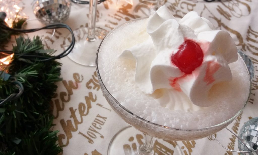 Close up of a champagne glass filled with eggnog, whipped cream and a cherry on a table cloth with holiday decoration.